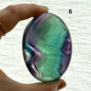 Puede incluir: Una piedra de fluorita pulida de forma ovalada. La piedra presenta una mezcla de colores, incluyendo verde, morado y azul. El n&uacute;mero "6" es visible en la esquina superior derecha. La piedra es sostenida por una mano.