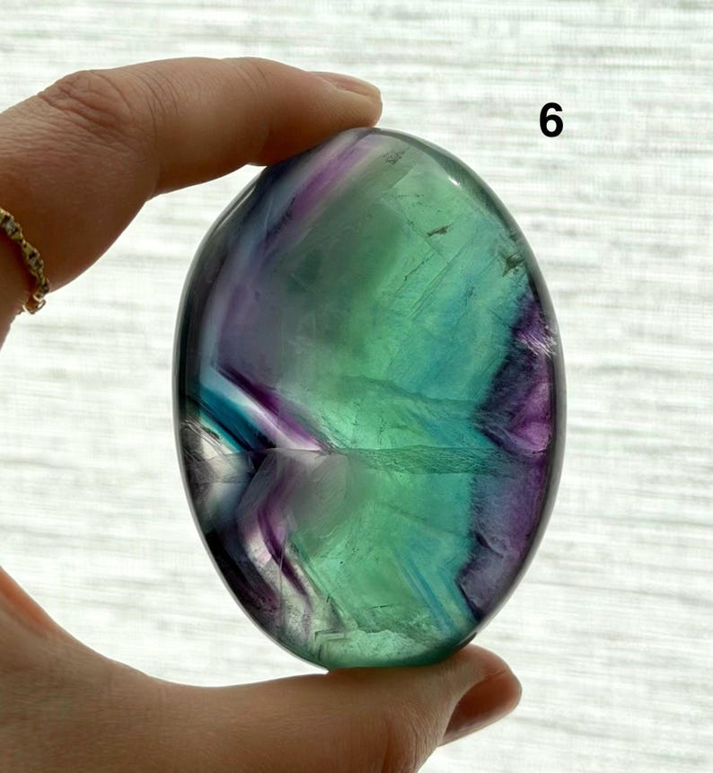 Puede incluir: Una piedra de fluorita pulida de forma ovalada. La piedra presenta una mezcla de colores, incluyendo verde, morado y azul. El n&uacute;mero "6" es visible en la esquina superior derecha. La piedra es sostenida por una mano.
