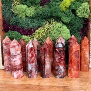 May include: A collection of red and white quartz crystal towers. The crystals are arranged in a row on a wooden surface, with a backdrop of green and purple moss. The crystals have a variety of patterns and textures.