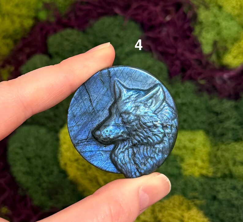 May include: A round, blue labradorite stone with a carved wolf profile. The stone has a polished finish and displays iridescent blue and gray hues. The number "4" is visible in the background. The stone is held between two fingers.