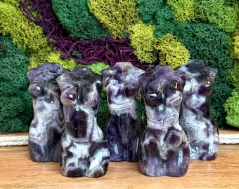 Puede incluir: Cinco tallas de cristal de amatista pulida con forma de torso humano. Cada pieza muestra variaciones naturales de bandas de color púrpura oscuro y blanco, con una superficie lisa y reflectante que resalta los patrones minerales únicos.