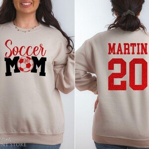Puede incluir: Sudadera beige con las palabras "Soccer Mom" en letras rojas y negras, con un gr&aacute;fico de bal&oacute;n de f&uacute;tbol. La parte trasera de la sudadera muestra el nombre "MARTIN" y el n&uacute;mero "20" en rojo.