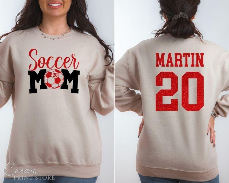 Puede incluir: Sudadera beige con las palabras "Soccer Mom" en letras rojas y negras, con un gr&aacute;fico de bal&oacute;n de f&uacute;tbol. La parte trasera de la sudadera muestra el nombre "MARTIN" y el n&uacute;mero "20" en rojo.