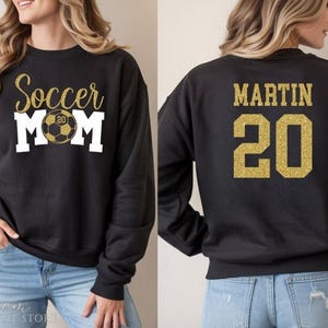 Puede incluir: Sudadera negra con texto dorado con purpurina que dice "Soccer Mom" con un gr&aacute;fico de bal&oacute;n de f&uacute;tbol. La parte trasera de la sudadera tiene el nombre "MARTIN" y el n&uacute;mero "20" en purpurina dorada.