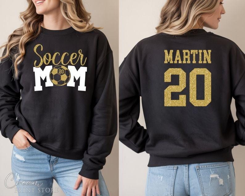 Puede incluir: Sudadera negra con texto dorado con purpurina que dice "Soccer Mom" con un gr&aacute;fico de bal&oacute;n de f&uacute;tbol. La parte trasera de la sudadera tiene el nombre "MARTIN" y el n&uacute;mero "20" en purpurina dorada.