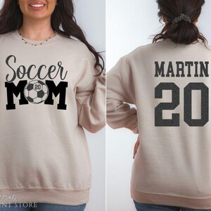 Puede incluir: Sudadera beige con el texto "Soccer Mom" en negro, con un gr&aacute;fico de bal&oacute;n de f&uacute;tbol con el n&uacute;mero "20". La parte trasera de la sudadera muestra el nombre "MARTIN" y el n&uacute;mero "20" en negro.