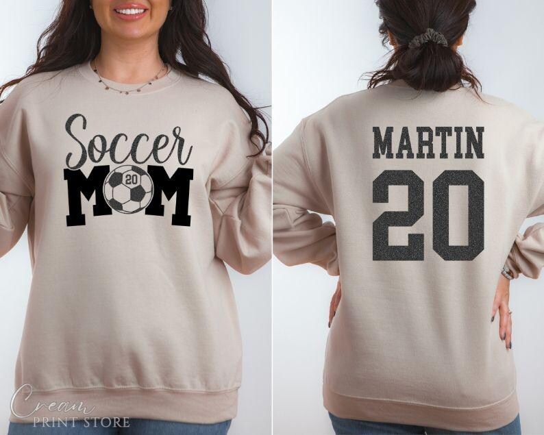Puede incluir: Sudadera beige con el texto "Soccer Mom" en negro, con un gr&aacute;fico de bal&oacute;n de f&uacute;tbol con el n&uacute;mero "20". La parte trasera de la sudadera muestra el nombre "MARTIN" y el n&uacute;mero "20" en negro.