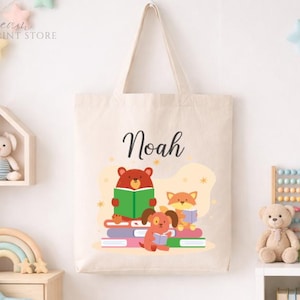May include: A beige tote bag with the name "Noah" in black script. The bag features a colorful illustration of a bear, fox, and dog reading books. The design is set against a light yellow background with star accents. The bag has a simple handle.
