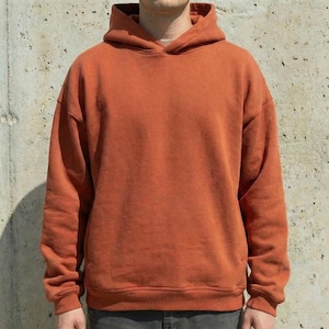 Puede incluir: Una sudadera con capucha de color naranja quemado con capucha con cordón. La sudadera tiene mangas largas y un ajuste relajado. La tela parece ser un material suave y de peso medio. La sudadera con capucha es de color liso sin gráficos ni patrones visibles.