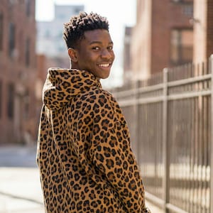 Pode incluir: Uma pessoa vestindo um moletom com capuz com estampa de leopardo marrom e preto. O moletom tem capuz e é feito de um material macio. A pessoa está sorrindo e olhando por cima do ombro.