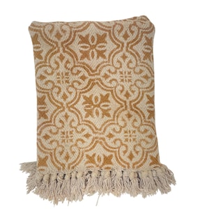 May include: A decorative cushion with a beige background and a repeating gold floral pattern. The cushion has a fringe trim at the bottom. The cushion is rectangular in shape.