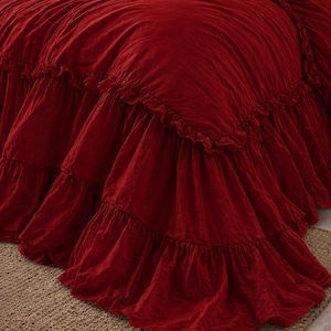 May include: A crimson bedspread with a frilled skirt. The bedding is a solid, deep red colour and appears to be made of a soft fabric. The frilled skirt adds a decorative element to the bed.