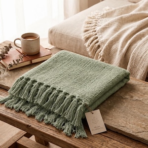 Puede incluir: Una manta tejida de color verde salvia con una superficie de tela texturizada y flecos largos en los bordes. Una pequeña etiqueta de papel rectangular con un logotipo de hoja y el texto Woven Comfort Co. está unida a la esquina de la manta.