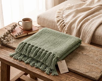 May include: A sage green woven throw blanket featuring a textured fabric surface and long fringe trim along the edges. A small rectangular paper tag with a leaf logo and the text Woven Comfort Co. is attached to the corner of the folded blanket.