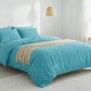 Puede incluir: Una cama con una funda nórdica turquesa y fundas de almohada a juego. Una manta beige con flecos está colocada sobre la cama. La cama está sobre una alfombra de color claro en una habitación luminosa. La estructura de la cama es de madera.