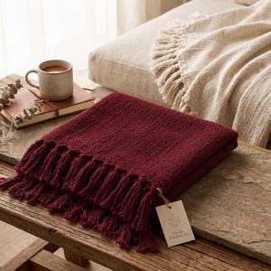 May include: A deep burgundy throw blanket with a textured, woven surface and long fringe trim on the edges. A small, rectangular beige paper tag is attached to the corner, featuring a line-art leaf icon and the text Woven Comfort Co.