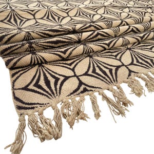 May include: Beige and black patterned textile with a floral design. The fabric has a repeating pattern of stylized flowers in black against a beige background. The edge is finished with beige fringe.