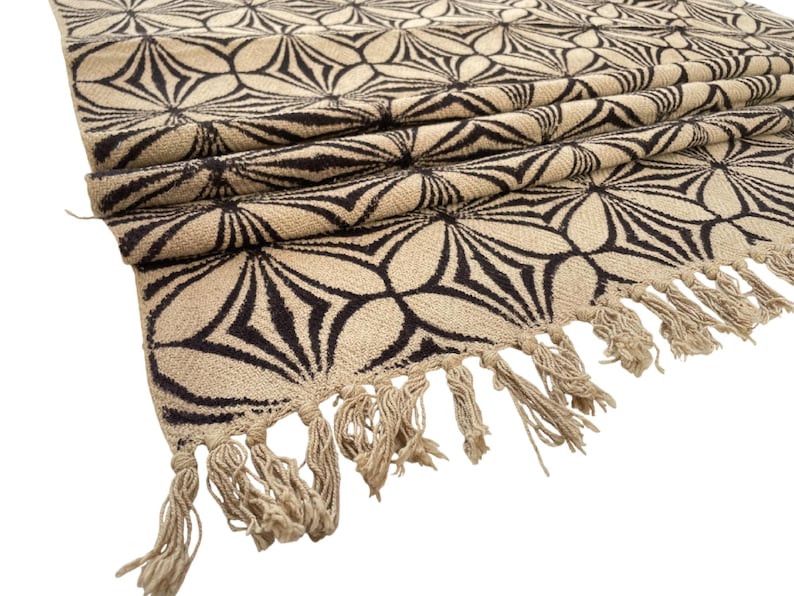 May include: Beige and black patterned textile with a floral design. The fabric has a repeating pattern of stylized flowers in black against a beige background. The edge is finished with beige fringe.
