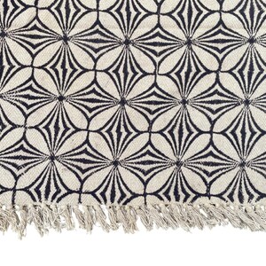 May include: Cream-colored rug with a repeating black floral pattern. The design features stylized flower shapes. The rug has a fringe along one edge. This rug is suitable for living rooms, bedrooms, or entryways.