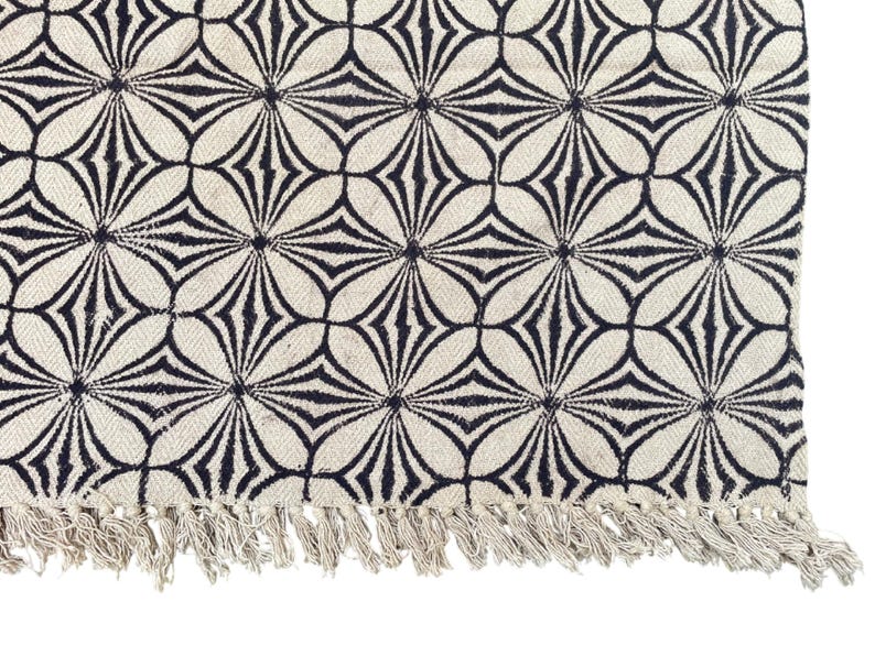 May include: Cream-colored rug with a repeating black floral pattern. The design features stylized flower shapes. The rug has a fringe along one edge. This rug is suitable for living rooms, bedrooms, or entryways.