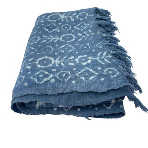 May include: Folded blue blanket with white geometric patterns and fringe trim. The blanket features a repeating design of circles, diamonds, and floral elements. The fabric appears to be a woven material.