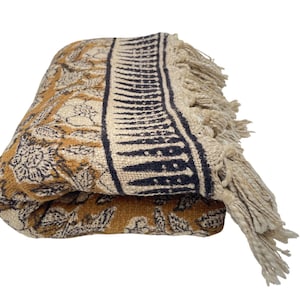 Könnte beinhalten: Gefaltete senffarbene Wurkdecke mit floralem Muster und cremefarbenem Rand mit geometrischem Design. Die Decke hat an einem Ende Fransen. Der Überwurf besteht aus einem weichen, gewebten Material.