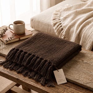 Puede incluir: Una manta tejida de color marrón oscuro con una superficie texturizada y flecos en los bordes. Una pequeña etiqueta rectangular blanca está unida a la esquina, con un dibujo lineal de una planta y el texto Woven Comfort Co. en tinta negra.