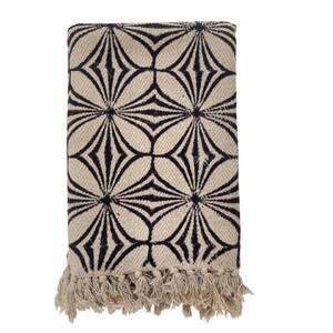May include: Folded beige throw blanket with a repeating black geometric floral pattern. The blanket has a fringed edge at the bottom. The design is a modern take on a classic pattern, suitable for a living room or bedroom.