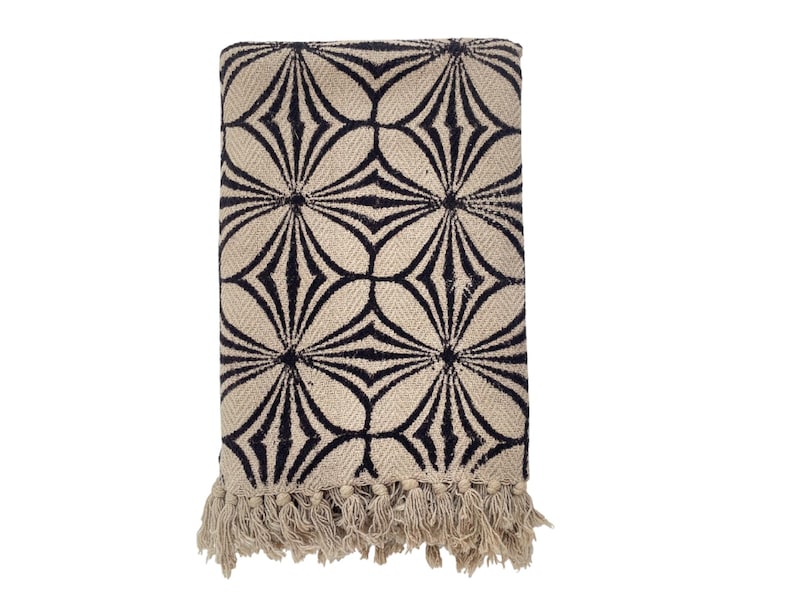 May include: Folded beige throw blanket with a repeating black geometric floral pattern. The blanket has a fringed edge at the bottom. The design is a modern take on a classic pattern, suitable for a living room or bedroom.
