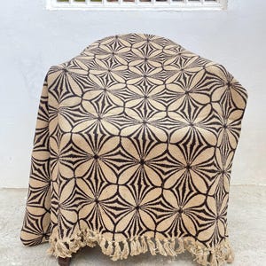 May include: A beige and brown patterned throw blanket draped over a chair. The blanket features a repeating floral design and a fringed edge. The neutral color palette makes it suitable for various home decor styles.