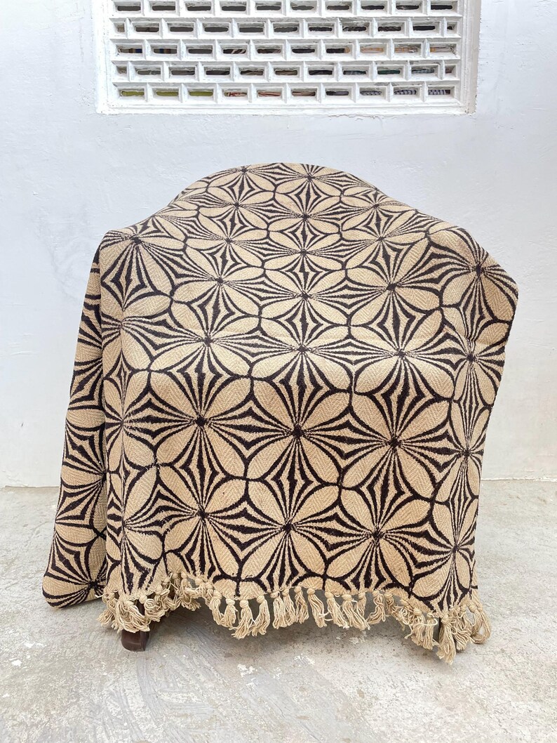 May include: A beige and brown patterned throw blanket draped over a chair. The blanket features a repeating floral design and a fringed edge. The neutral color palette makes it suitable for various home decor styles.