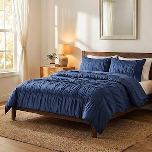 May include: A navy blue bedspread and pillow set on a wooden bed frame. The bedding has a textured, gathered design. The room features a neutral color palette with a jute rug and a bedside table with a lamp.