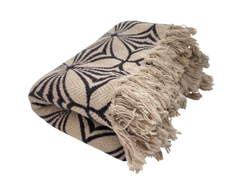 May include: Folded beige throw blanket with a black floral pattern. The blanket has a fringed edge. The pattern is a repeating design of stylized flower shapes. The blanket is made of a woven fabric.