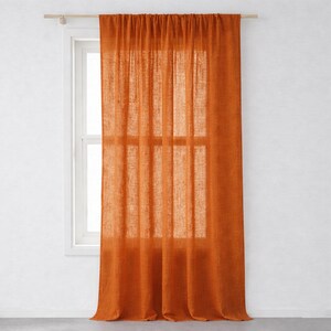 Op de afbeelding: Een enkele, verbrande oranje linnen gordijn hangt voor een wit raam. Het gordijn is opgehangen aan een houten roede. Het raam is verdeeld in meerdere ruiten. Het gordijn is gemaakt van een semi-transparante stof die wat licht doorlaat.
