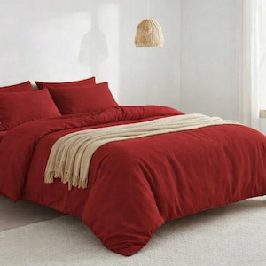 Puede incluir: Una cama con una funda nórdica de color rojo intenso y fundas de almohada a juego. Una manta beige con flecos está colocada sobre la cama. La cama está sobre una alfombra de color claro. También se ve una pequeña mesita de noche de madera y una lámpara colgante tejida.