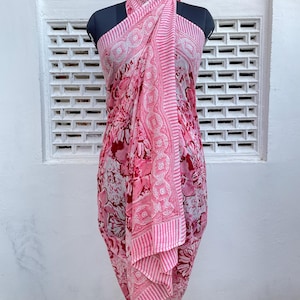 Op de afbeelding: Een roze en wit patroon sarong gedrapeerd op een mannequin. De sarong heeft een bloemenpatroon met een gestreepte rand. De stof is lichtgewicht en lijkt van katoen te zijn. De sarong is om de torso en benen van de mannequin gewikkeld.