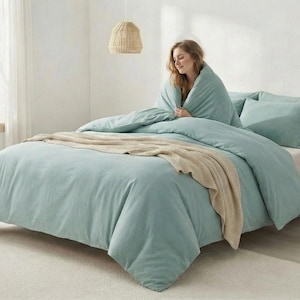 May include: A bed with light blue bedding, including a duvet cover and pillows. A beige throw blanket is draped across the bed. A person is wrapped in the duvet. A small side table with a cup and dried flowers is next to the bed.