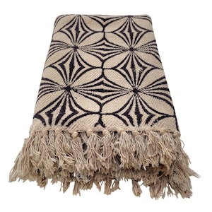 May include: A beige throw blanket with a repeating black geometric pattern. The blanket has a fringe trim along the bottom edge. The pattern is a series of connected starburst shapes.