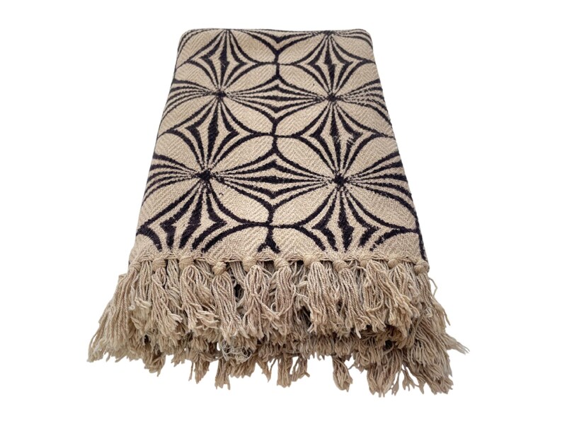 May include: A beige throw blanket with a repeating black geometric pattern. The blanket has a fringe trim along the bottom edge. The pattern is a series of connected starburst shapes.