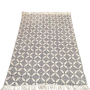 May include: A rectangular rug with a repeating floral pattern in black and off-white. The rug has a fringe along the short edges. The design features a series of overlapping, stylized flower shapes.
