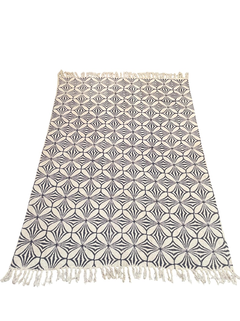 May include: A rectangular rug with a repeating floral pattern in black and off-white. The rug has a fringe along the short edges. The design features a series of overlapping, stylized flower shapes.
