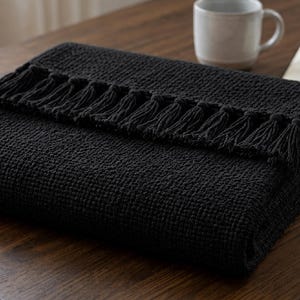 May include: A folded black throw blanket featuring a textured waffle weave pattern and a row of decorative fringe along the top edge. The blanket is rectangular in shape and displays a consistent dark charcoal color throughout its entire surface.
