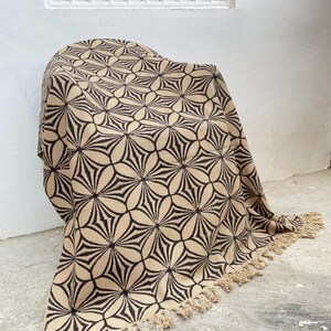 May include: A beige and black patterned throw blanket with a floral design. The blanket is draped over a chair and has a fringe trim along the bottom edge. The pattern is a repeating geometric design.