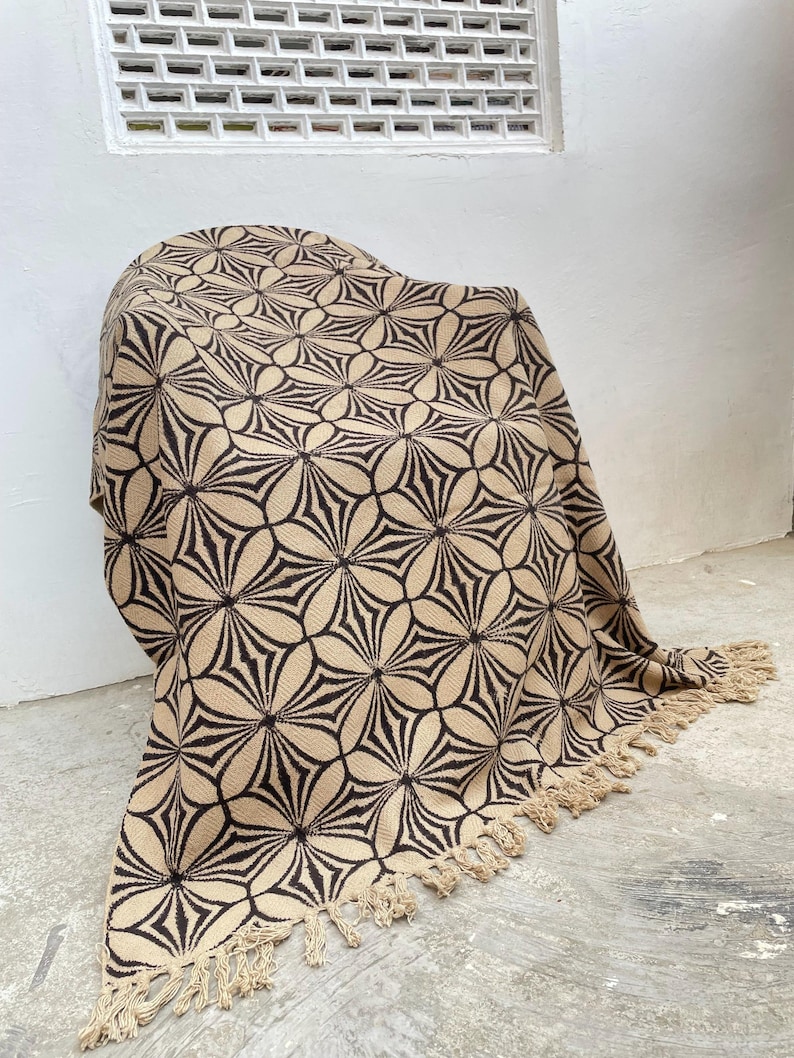 May include: A beige and black patterned throw blanket with a floral design. The blanket is draped over a chair and has a fringe trim along the bottom edge. The pattern is a repeating geometric design.
