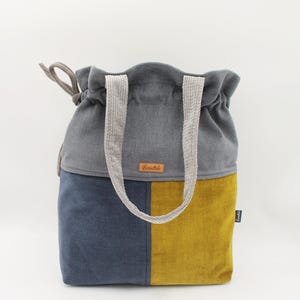 Może przedstawiać: Dwukolorowa torba typu tote z zapięciem na sznurek. Górna część jest szara, a dolna podzielona na granatowe i musztardowe panele. Torba ma szare uchwyty i małą etykietę z napisem "Scisttola".