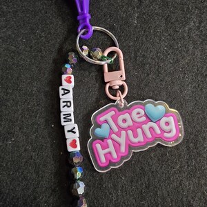 May include: A keychain with a purple clip, a pink clasp, and a charm that says "Tae Hyung" with blue hearts. The keychain also has a beaded strand with the word "ARMY" spelled out with red hearts.