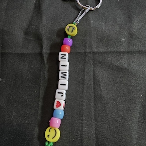 May include: A beaded keychain with the name "JIMIN" spelled out in white letter beads. The keychain features colorful beads, including yellow smiley faces, hearts, and a green tassel. It is attached to a silver keyring.