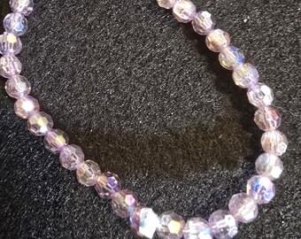 May include: A bracelet made of faceted, light purple beads. The beads are strung together to form a flexible band. The bracelet is displayed on a dark, textured surface. The beads have a shimmering, iridescent quality.