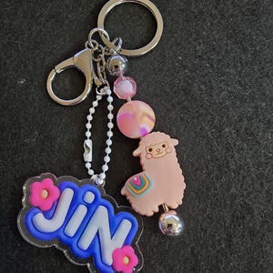 May include: A silver keychain with a lobster clasp, key ring, and beaded accents. The keychain features a pink llama charm with a rainbow design and a clear acrylic charm with the name "JIN" in blue and pink.