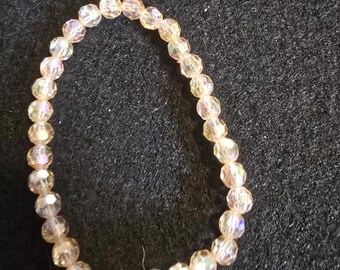 May include: A delicate bracelet composed of faceted, iridescent beads in shades of peach and cream. The beads are strung together, forming a circular shape. The bracelet is set against a dark background.
